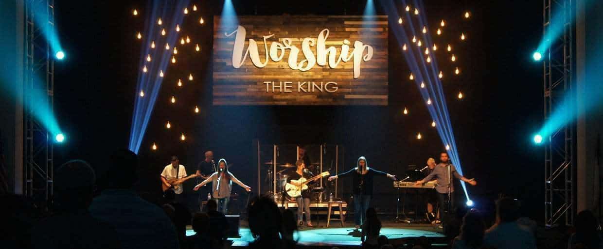 An eight-member praise and worship band performs on a darkened stage. The backdrop features hanging lightbulbs and a sign that reads "worship the king."
