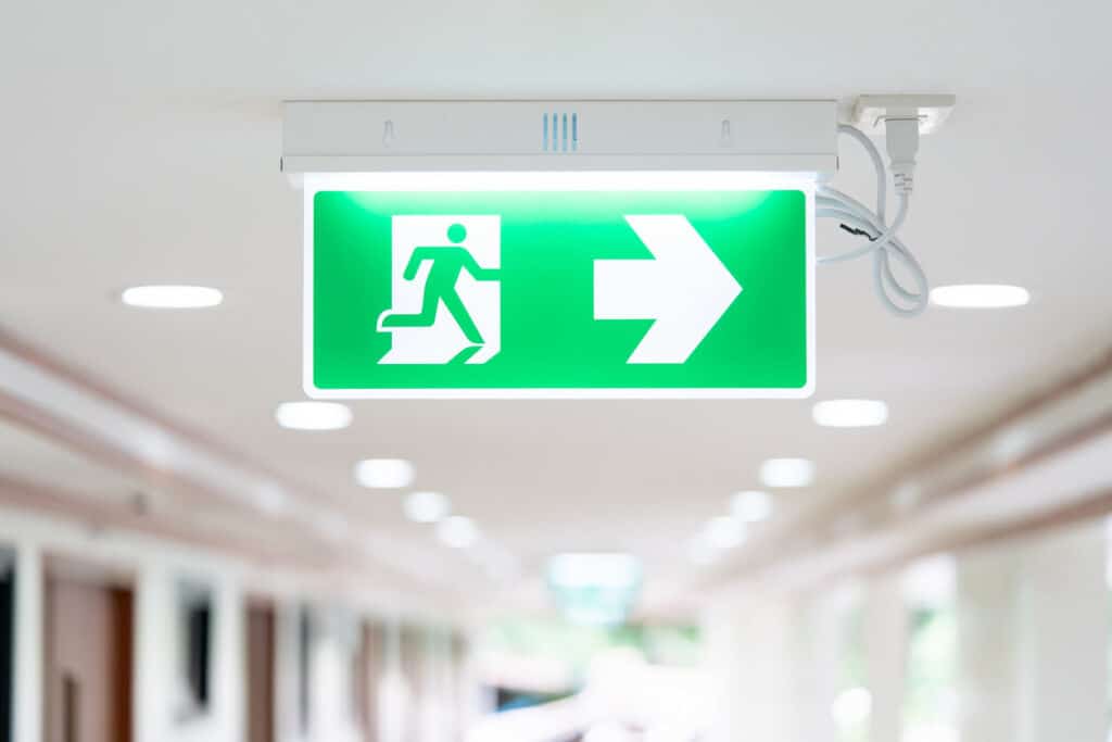 Wayfinding signage mounted to the ceiling of a hospital corridor showing a person exiting through a doorway and a right directional arrow.