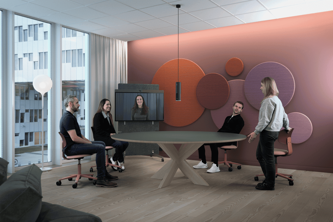 A team of four employees taking a video conference call in a meeting room. Three are seated around a round table and the fourth pulls out a chair to sit down.