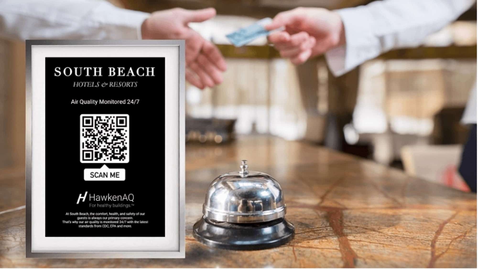 A sign displayed at a hotel check in desk featuring a QR code. A bell sits beside the sign while one person hands a credit card to another in the background.