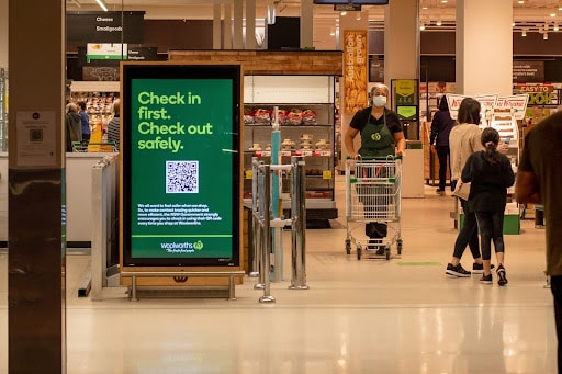 Digital QR code signage displayed within a grocery store entrance, surrounded by shoppers and employees.