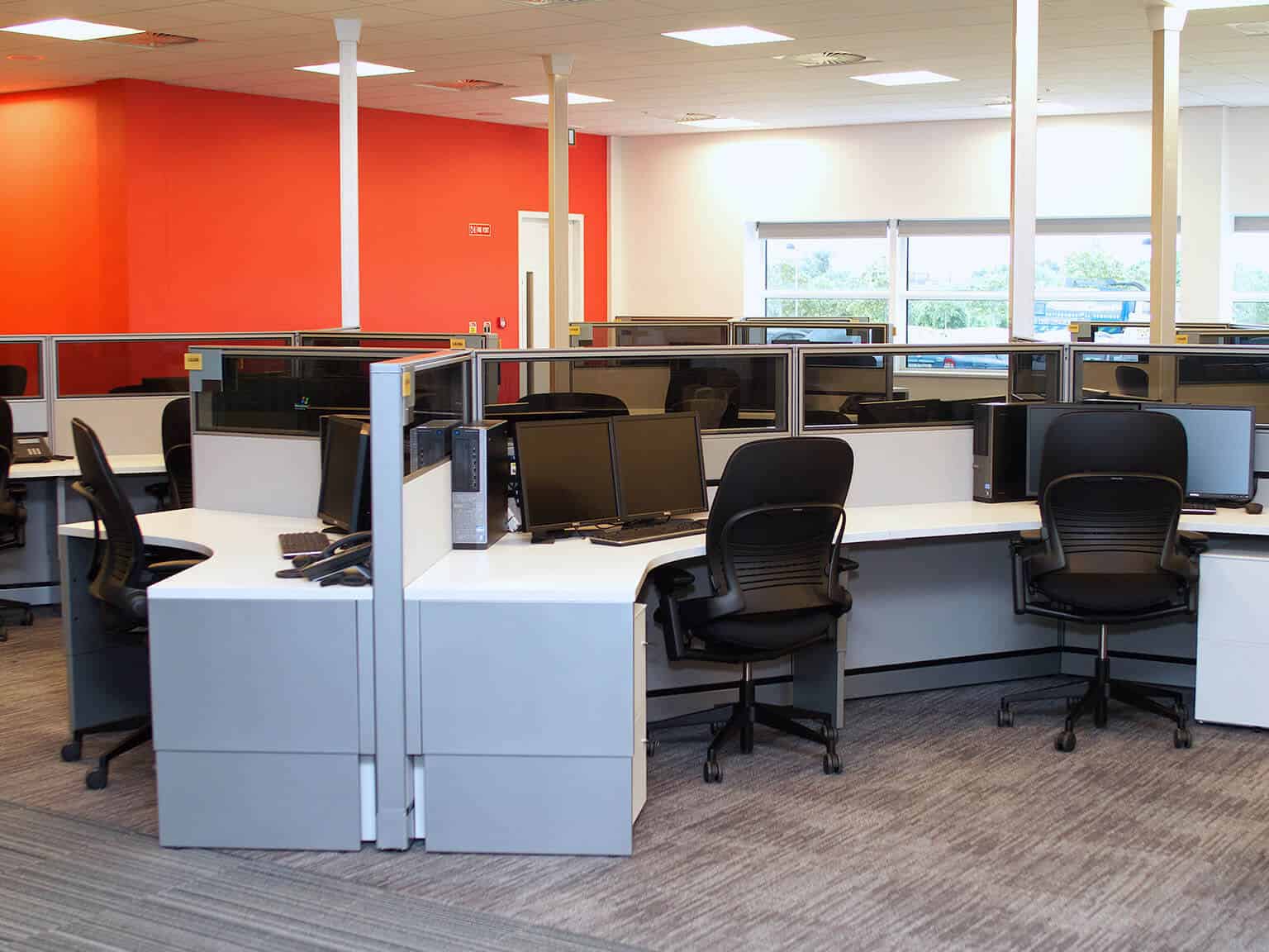 Office space with red and white walls, gray carpet, and cubicles. Each cubicle has two chairs and two monitors.