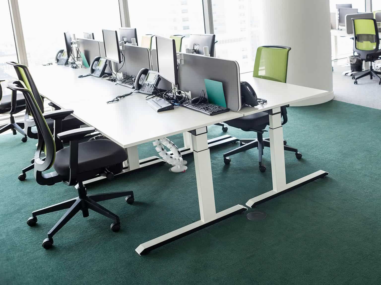 Long table on a green carpet with computer monitors, keyboards and phones. Green chairs sit at the table.