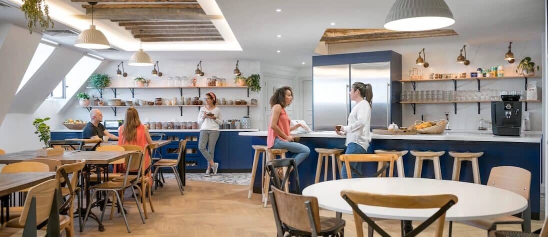 An employee break room in the Paris Airbnb office. The room has a wood floor and blue counters. Shelves line the white walls and are filled with glassware and plants. Some employees are seated at a table while others have a conversation in the kitchen area. Another employee stands in the background holding a bowl. 
