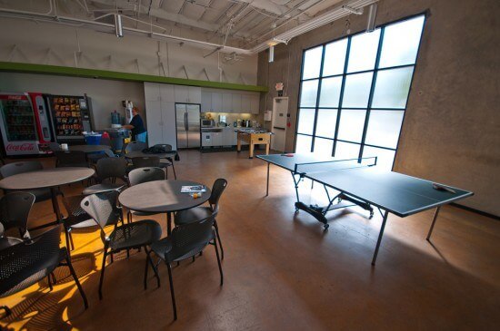 An employee break room at the Apple headquarters in Cupertino, California. The room has wooden floors, a large multi-pane window on the right wall, and an exposed ceiling. In the background is a kitchen area and two vending machines. Tables and chairs as well as a pin pong table are in the foreground.