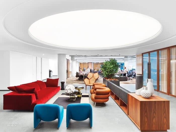 An open gathering space in Dropbox’s modern San Francisco office with white walls and a concrete floor. A large red couch and eclectic assortment of chairs make up a seating area that’s anchored by a wood-paneled storage shelf. 