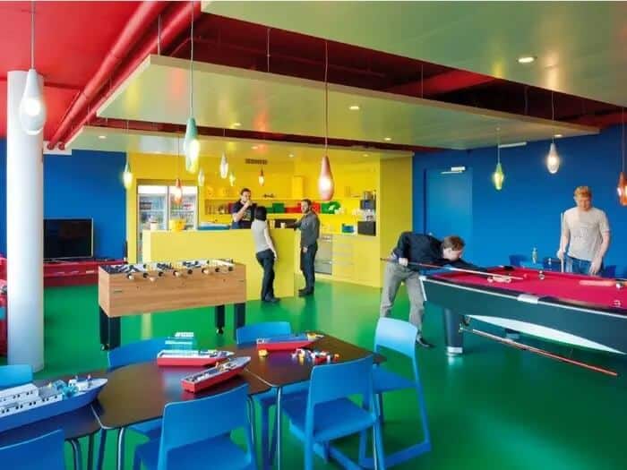 A colorful employee break room at Google’s Zurich office with blue and yellow walls, a green floor, and red ceiling. Some employees are standing in the kitchen area in the background while others play a game of pool.