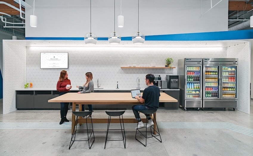 An open common area at LinkedIn headquarters with a grey floor and exposed ceiling. In the background is a kitchen area with white walls, a coffee bar, and three fully stocked refrigerators with glass doors. In the foreground, an employee sits at a communal table with their laptop while two other employees have a conversation. 