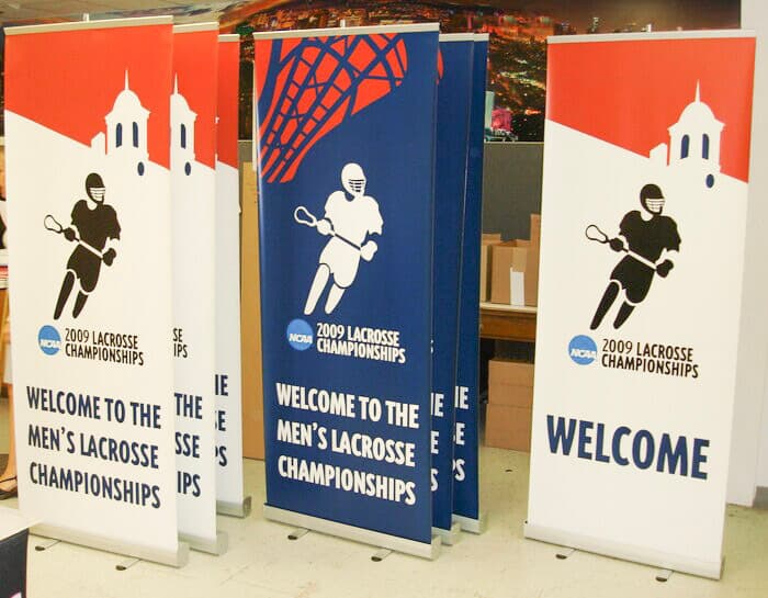 A display of seven retractable banners in navy blue, white and orange stand next to each other welcoming spectators to the NCAA Men’s Lacrosse Championship. There is an illustration of a lacrosse player on each banner.