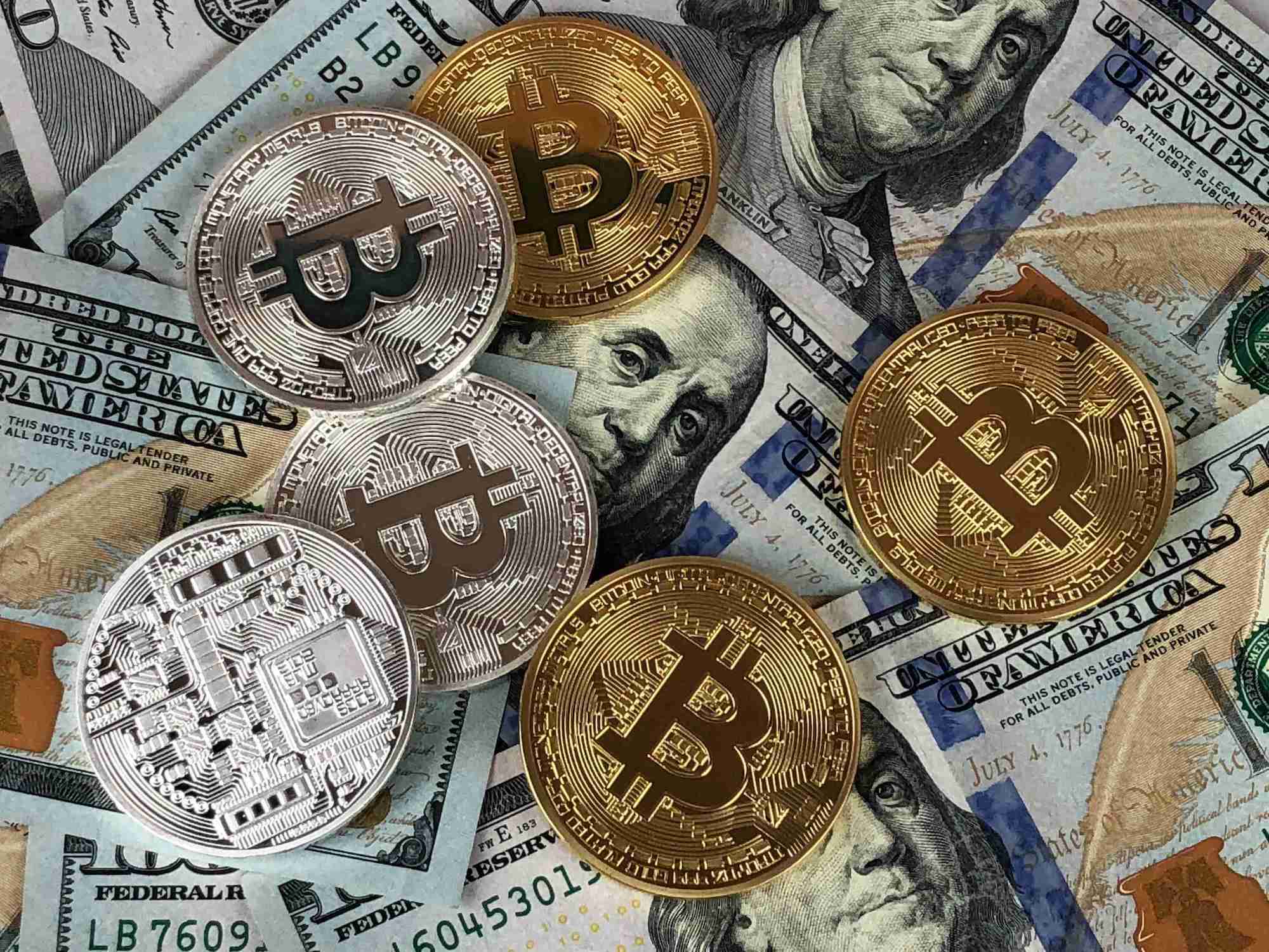 Six-gold-and-silver-coins-featuring-the-Bitcoin-symbol-lie-on-top-of-an-arrangement-of-100-dollar-bills.