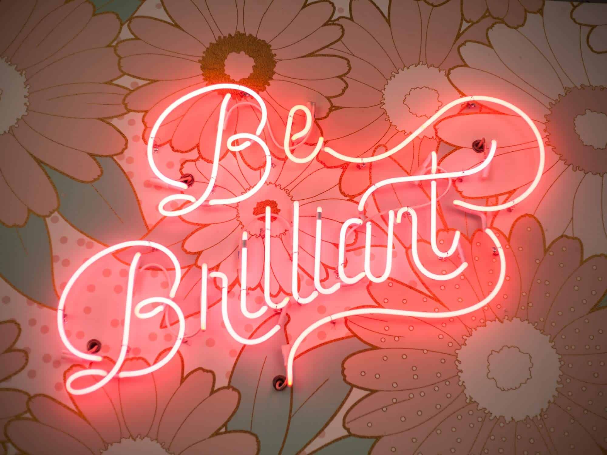 A-pink-neon-sign-that-reads-"Be-Brilliant"-in-looping-text-mounted-to-a-wall-with-floral-wallpaper.