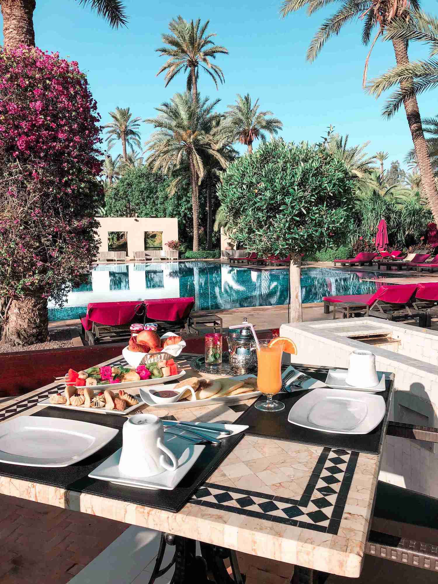 An-outdoor-pool-scene-featuring-a-table-set-for-two.-Lounge-chairs-are-placed-along-the-edge-of-the-pool-and-palm-trees-grow-in-the-background.