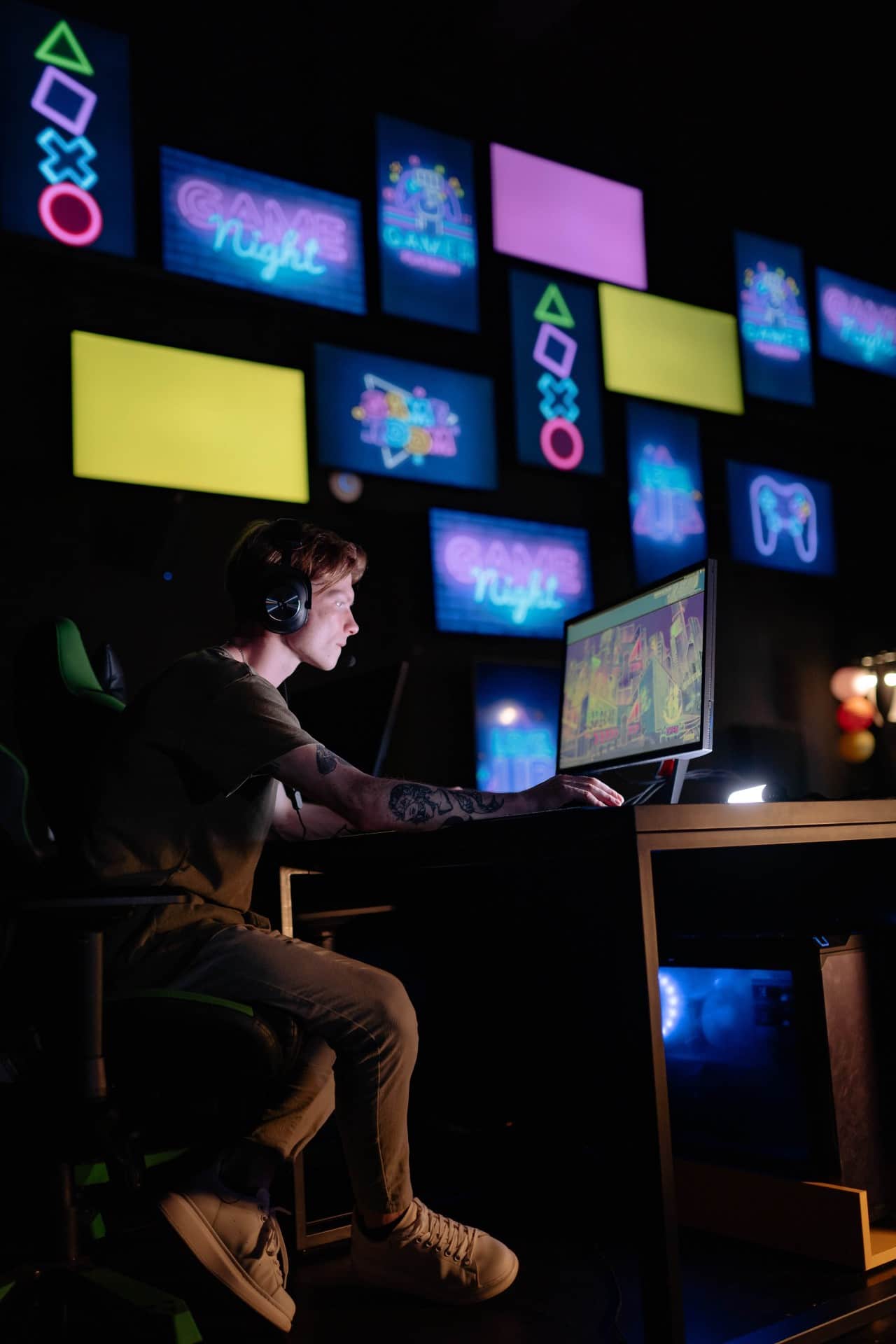 A man playing a PC video game with multi-colored neon signs on the wall next to him