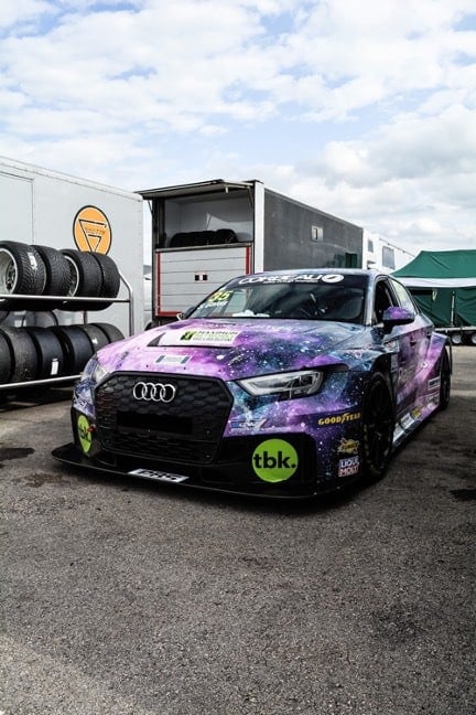 An Audi sits low to the ground adorned with a blue and purple galaxy themed car wrap with white-specks as stars. Sponsoring businesses are represented with stickers on the hood and front bumper areas.