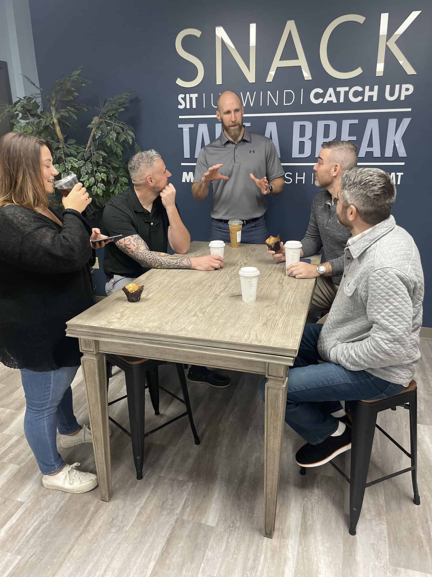 Five people are gathered around a table. One person stands at the head of the table and is speaking while the rest listen. Behind them is a dark blue wall with silver, white and light blue lettering.