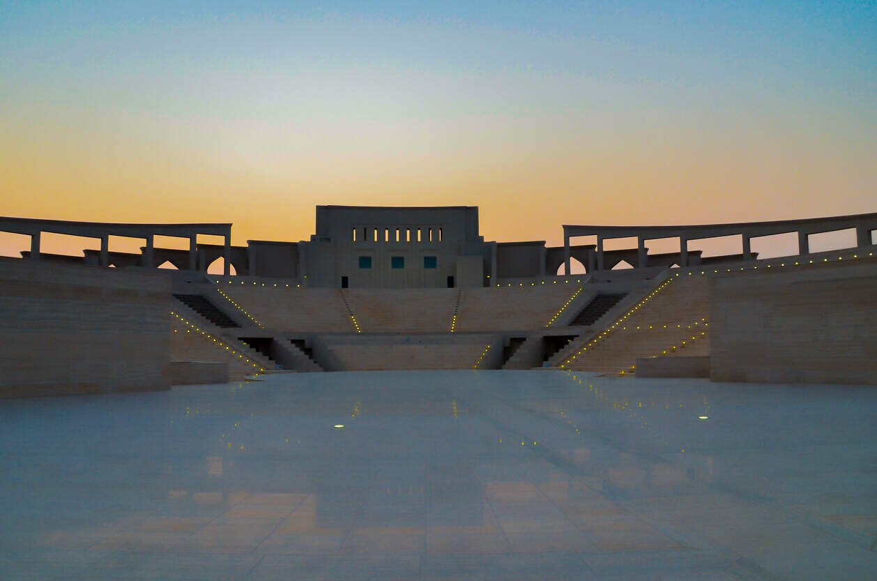 Modern amphitheater at sunset as seen from the stage. In the distance, floor lights illuminate the stadium-style seating area while terraces decorate the concourse.