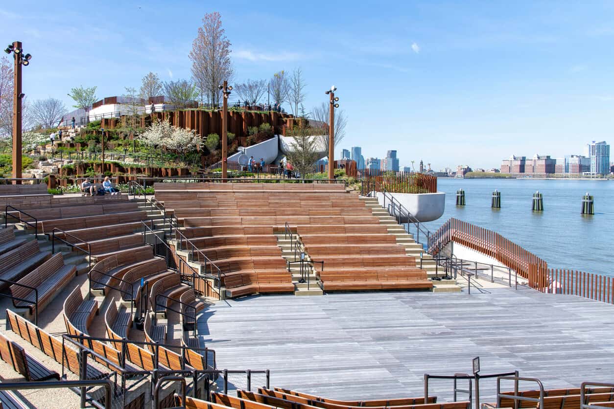 A waterfront amphitheater shown from the side. Wooden benches surround a round made from weathered wood planks. In the background are stepped gardens and a cityscape in the distance.