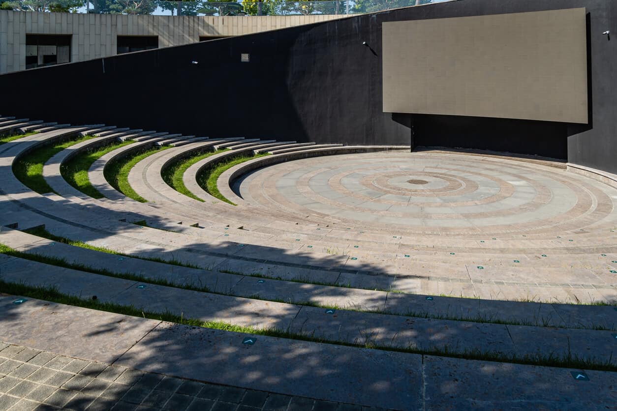 A stone amphitheater shown from the back row. Grass is planted in each row for comfortable seating. A large, blank screen is set up in the tiled round.