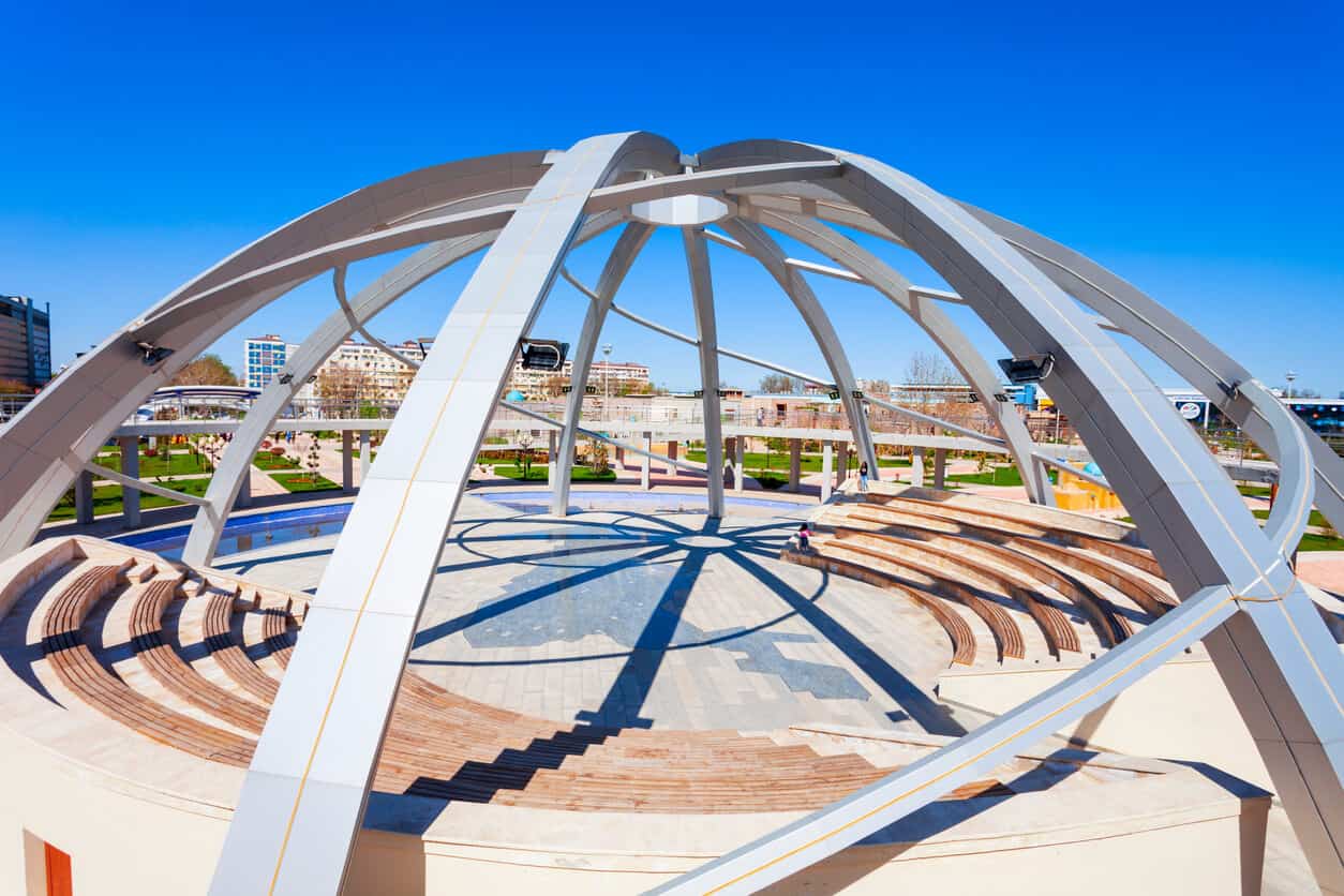 Stone and wood amphitheater with a planked round stage area. Arched metal beams create an open dome that covers the space. A manicured park area and buildings can be seen in the background.