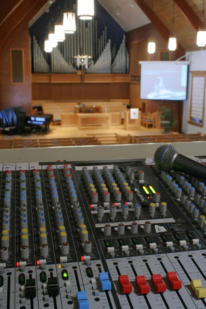 Close-up of a sound mixing board featuring red, yellow, and blue controls. A microphone rests on top of the board, and in the background is a church sanctuary.