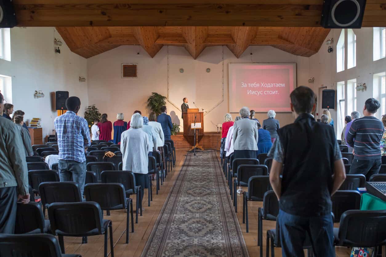 A church service in a room with a wood floor and ceiling and white walls. Two sections of seating are divided by a runner carpet. At the front, the pastor reads from a projector screen.