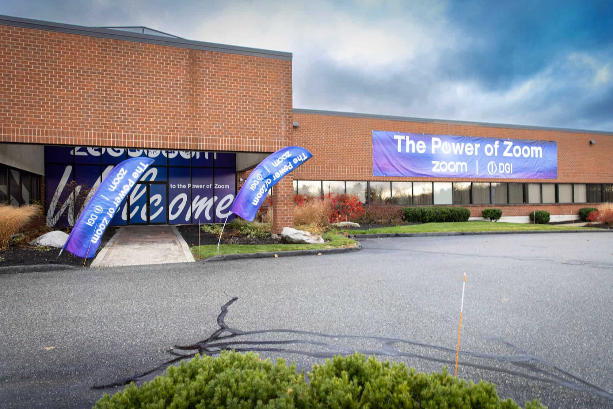 A brick building front featuring trade show signage. Blue banners with white writing are set up on the building, its entrance and in the lawn on either side of the walkway leading up to the front doors.