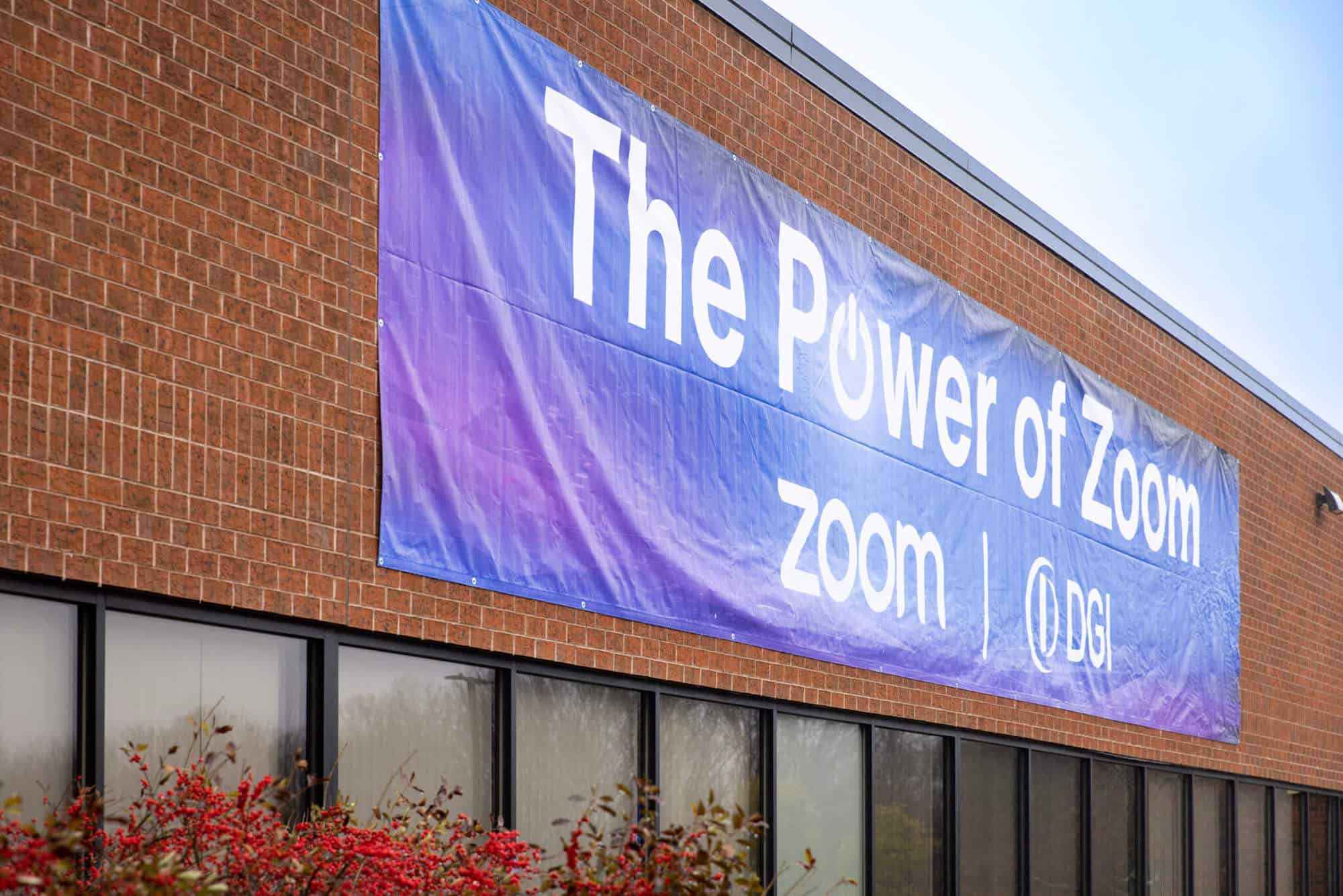 A blue trade show banner with white writing hanging above a row of windows on a brick building. The writing reads "The Power of Zoom."