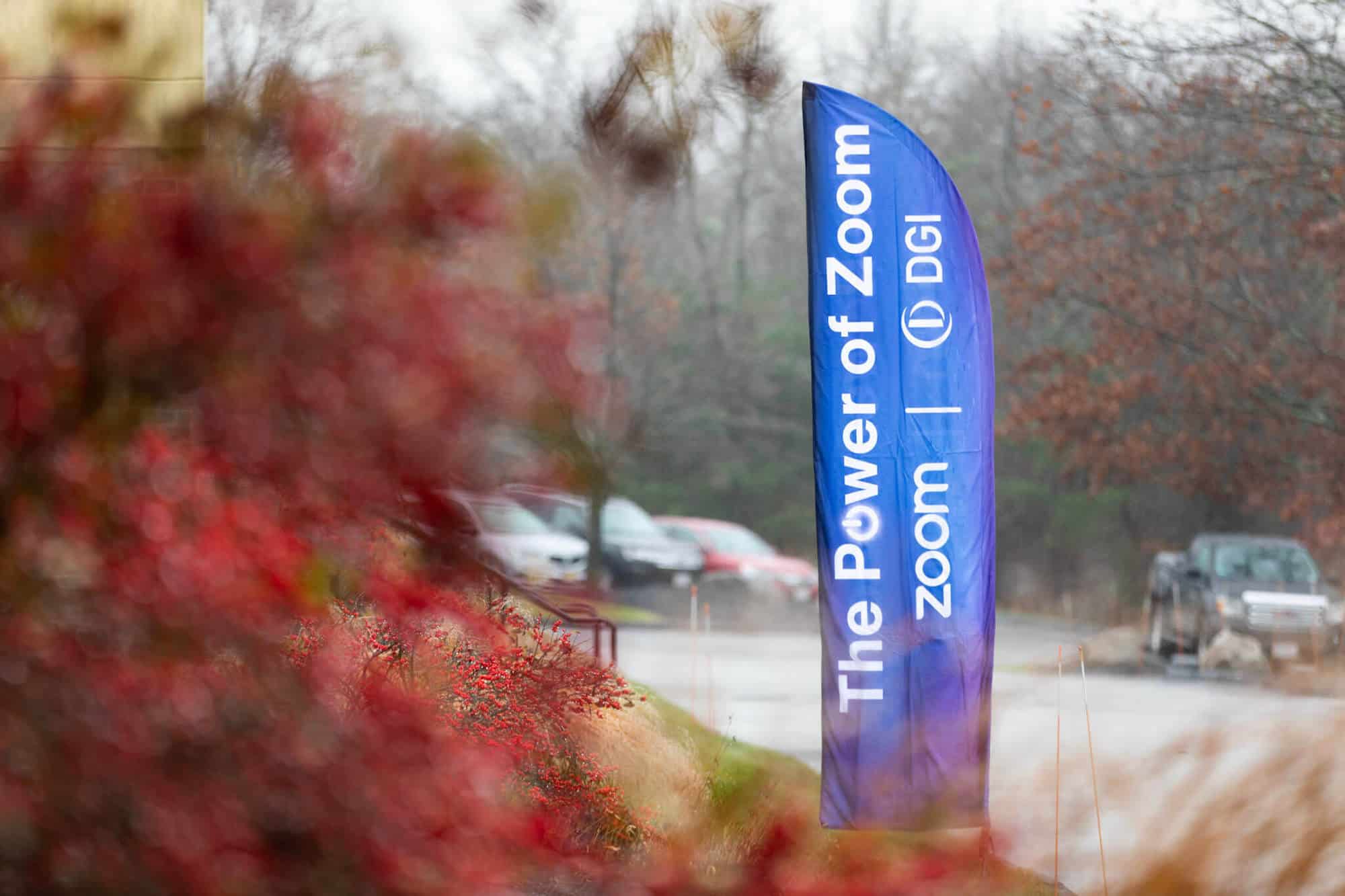 A blue banner with white writing that reads "The Power of Zoom," set up in a parking lot. A bush with red leaves is in the foreground.
