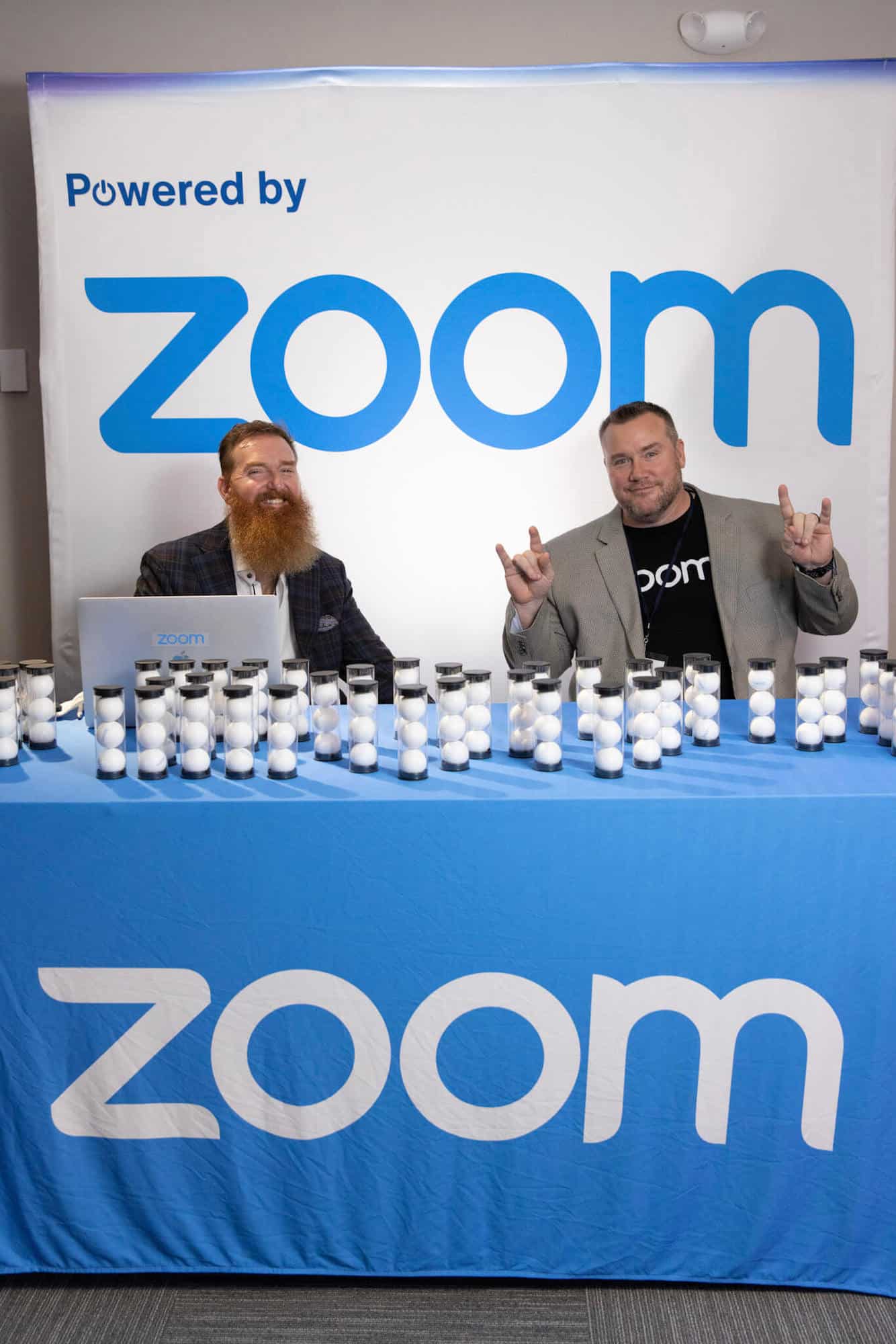 Two people sit at a trade show table covered in a blue cloth with white writing. The booth backdrop is white with blue writing that says "Powered by Zoom."