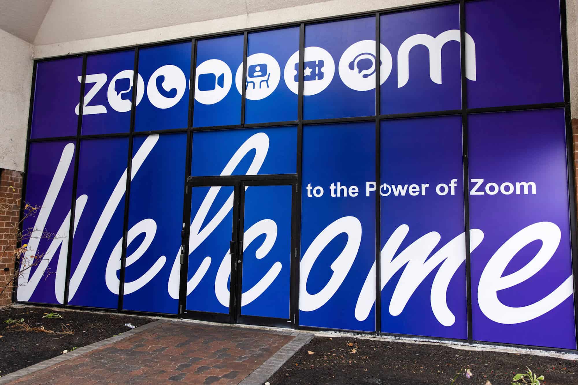 A close-up view of blue cling film trade show signage applied to the entrance to a brick building. White writing reads "Welcome to the Power of Zoom."
