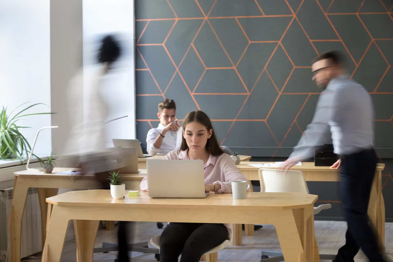 An open flexible workspace with with wooden tables. In the foreground, a woman sits and works at a laptop. Behind her, a man sits and looks at a piece of paper while two other employees walk by.