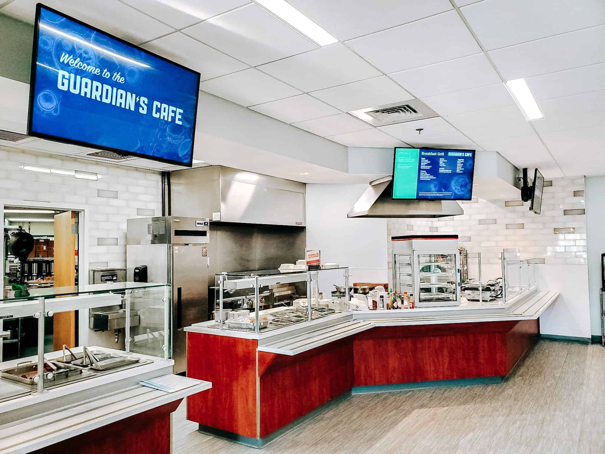 A cafeteria-style restaurant with food counters and a grill area. Digital signage displaying the name of the restaurant (Guardian's Cafe) and a menu are mounted on the ceiling.