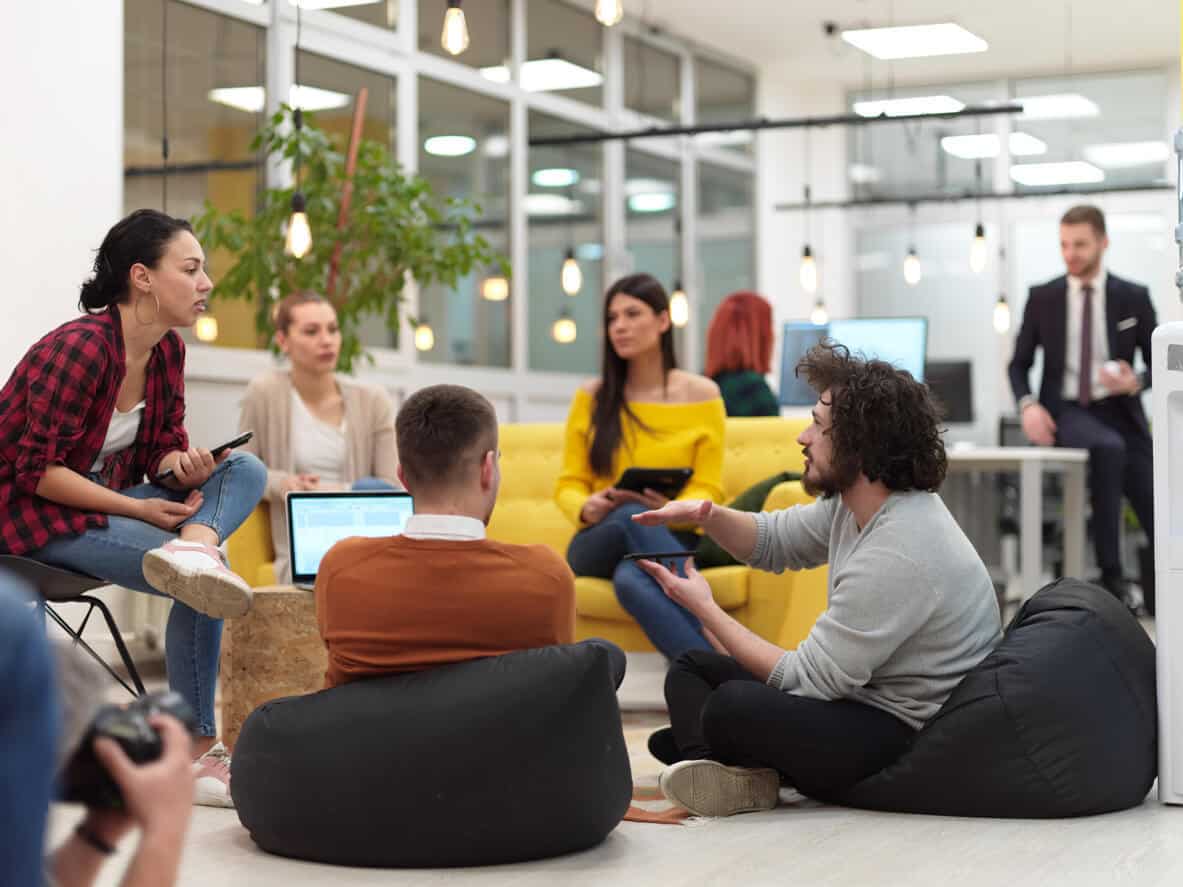 An open flexible workspace where three people are having a conversation while seated. Two men sit on bean bags while a woman sits on a chair. In the background two other women sit on a yellow couch.