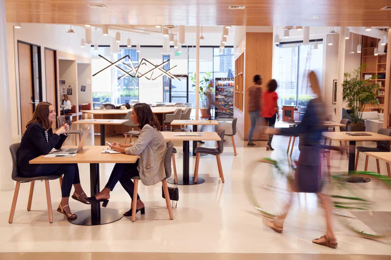A busy office cafeteria seating area and flexible workspace. Two women have a conversation while seated at a table. To the right, another woman passes through while walking a bike.