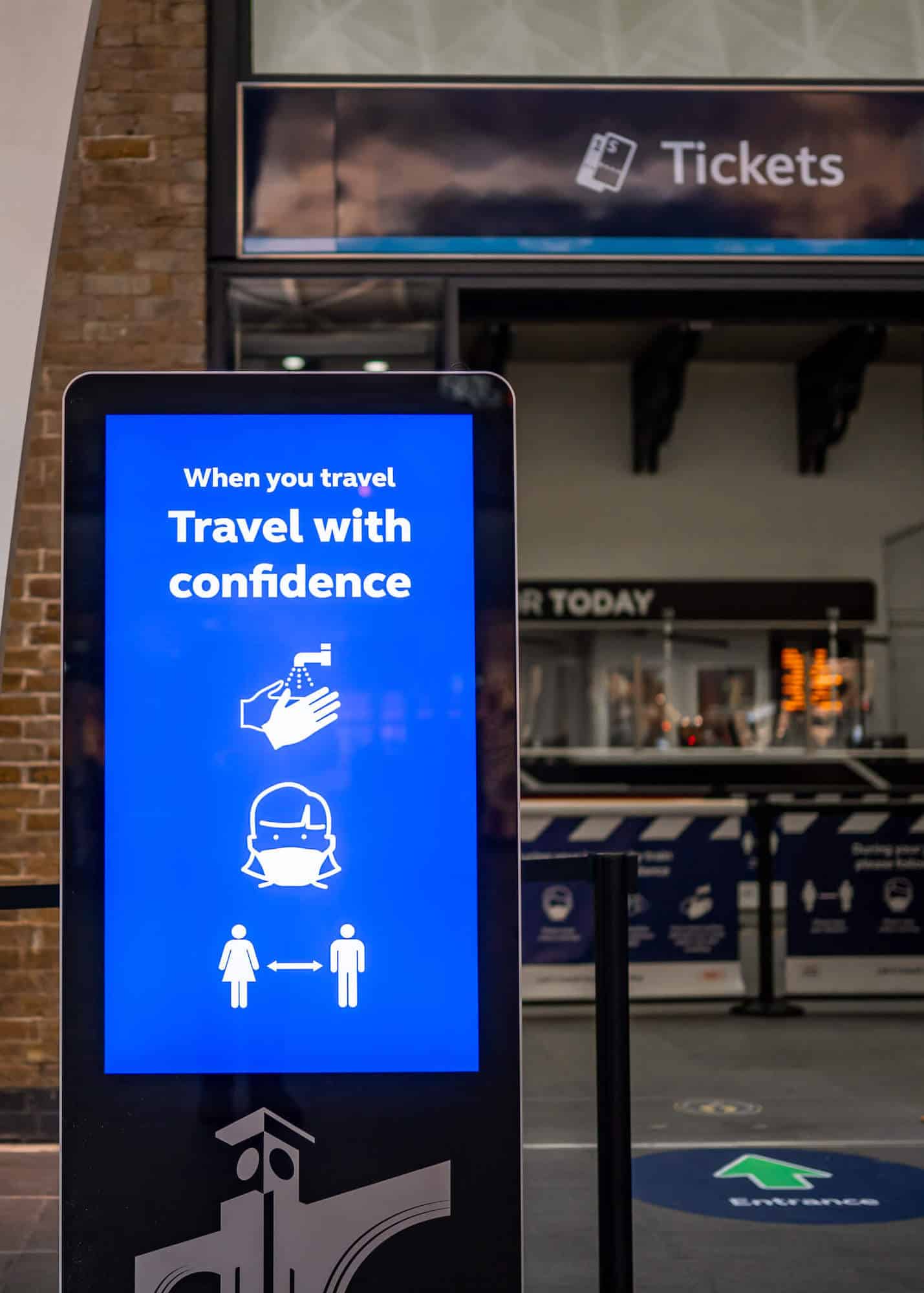 A digital sign located outside of the ticket office of a travel station. The sign reads "When you travel, travel with confidence" and displays white icons for hand washing, mask wearing and social distancing on a blue background.
