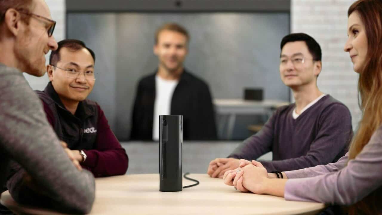 Four employees gathered around a table featuring video conferencing equipment. A fifth, remote meeting participant is on the screen in the background.