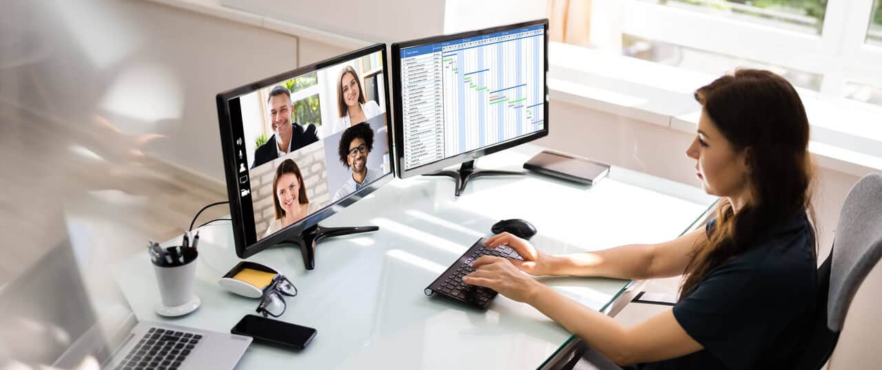 An employee attending a video conference call from their desk. There are two monitors on the desk, one shows remote meeting participants while the other features a spreadsheet.