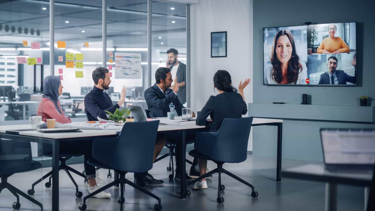 A conference room in a modern office with four people seated around a table. A screen at the front of the room shows three remote meeting participants.