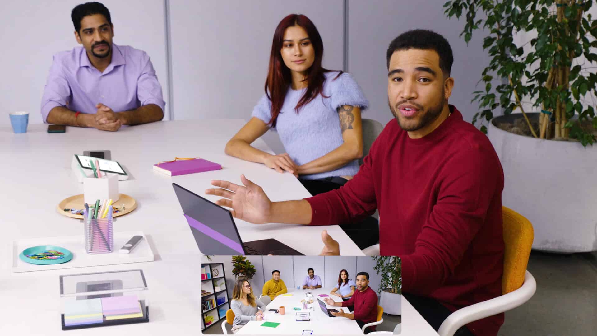 A close-up of three in-office meeting participants from the perspective of a webcam. An inset shows a wide-angle perspective of the conference room that includes all five participants.