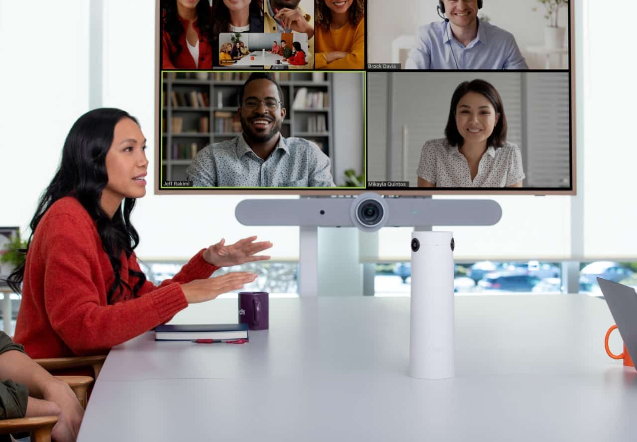 An in-office employee speaks while seated at a table during a video conference call. In the background is a monitor displaying remote participants.