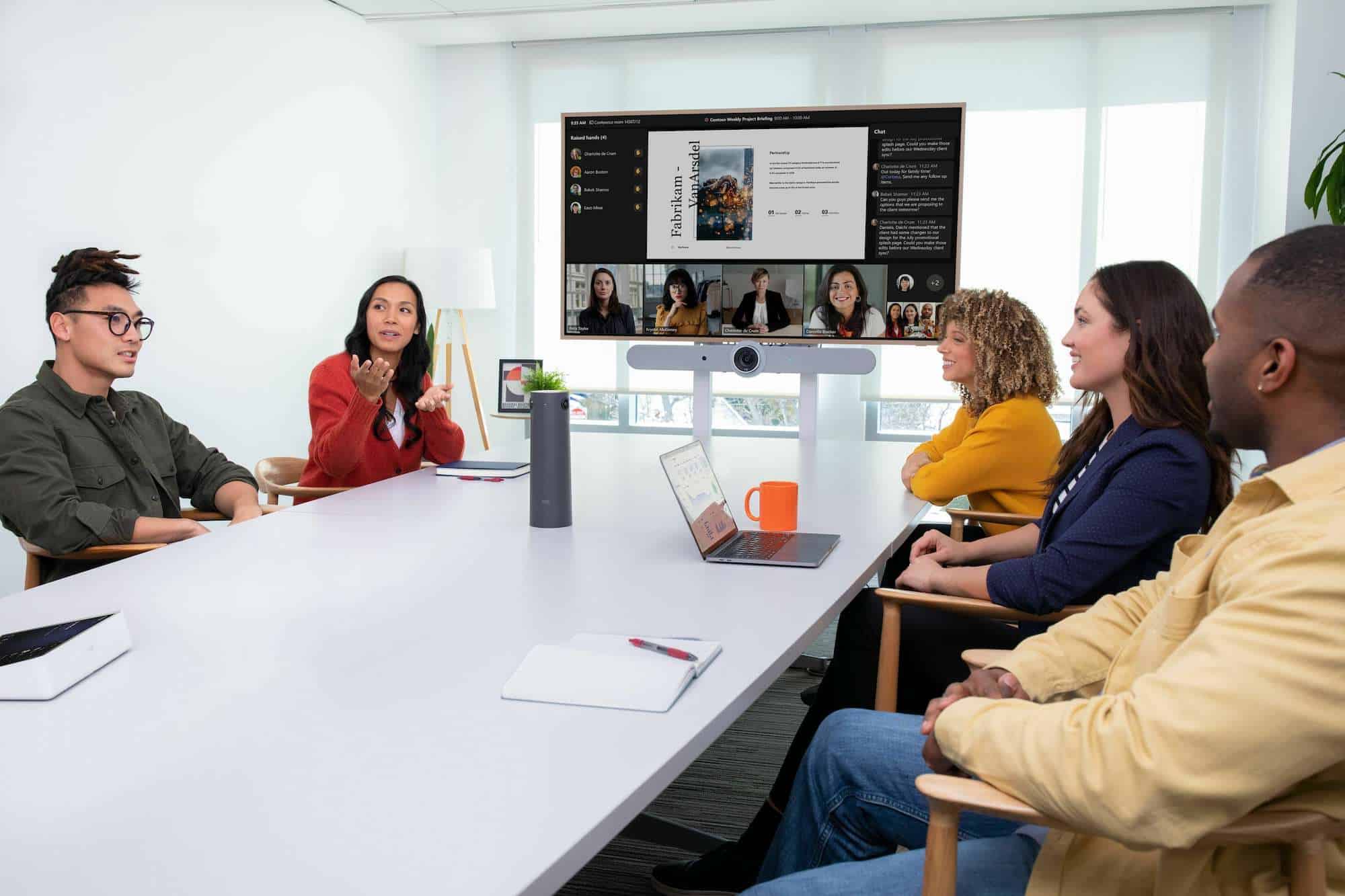 Five people participate in a virtual meeting held in a conference room with white walls and teams rooms equipment. At the head of the room a screen displays remote attendees and a presentation.