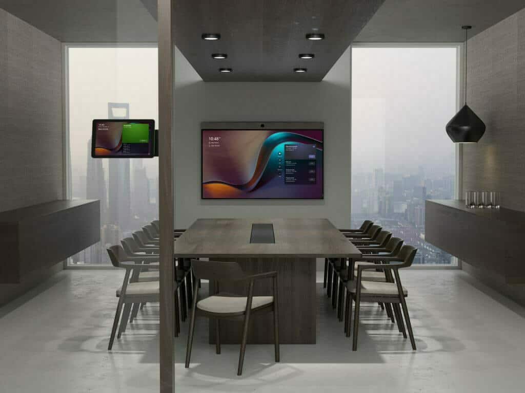 Teams rooms hardware is pictured in an empty conference room with gray walls, table, and chairs. A screen displaying virtual meeting software is mounted to the far wall, while a room reservation system is installed by the door.